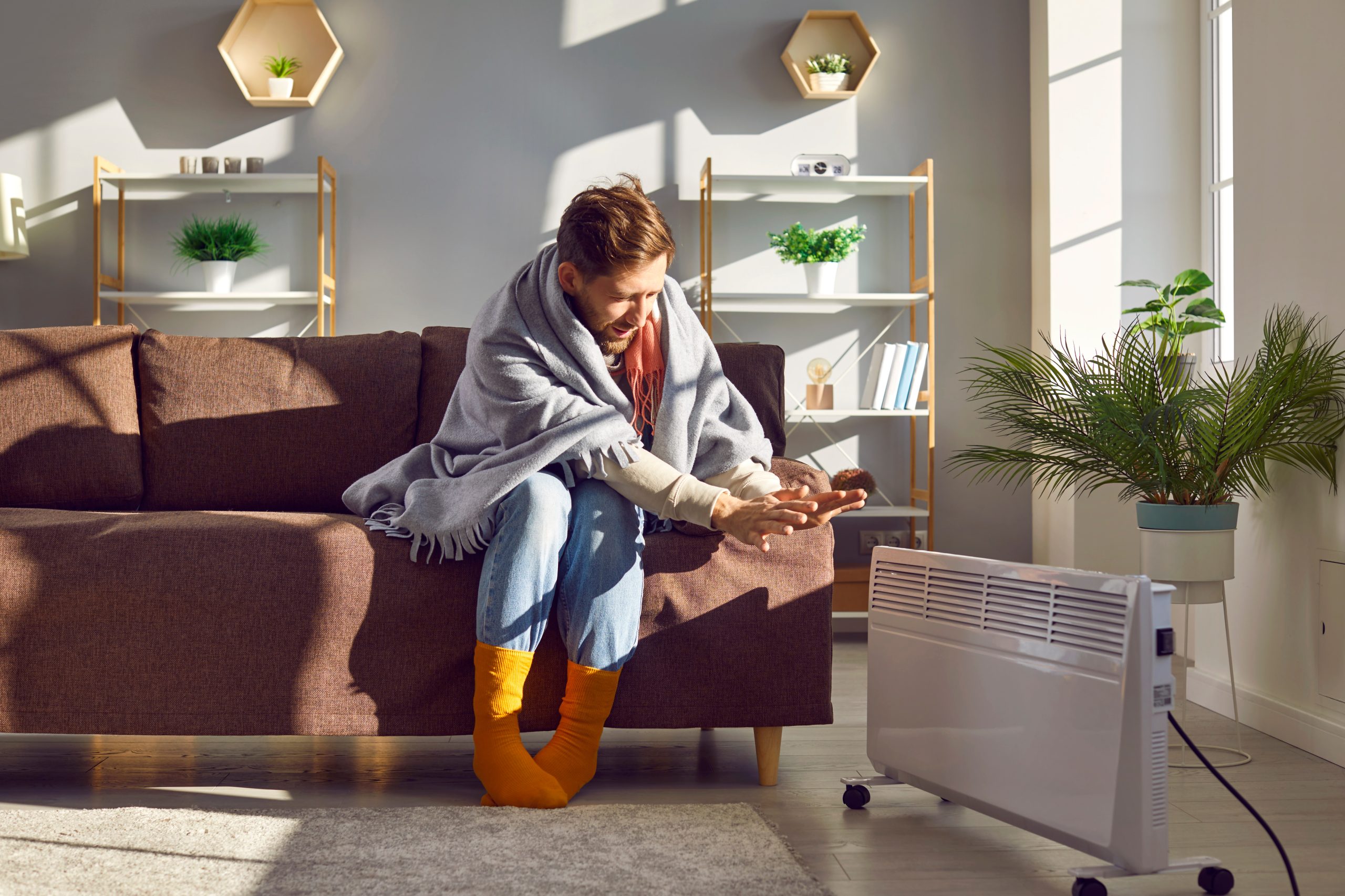 Man in socks and blanket warms his hands by a heater in a cold room at home in winter guy next to heater